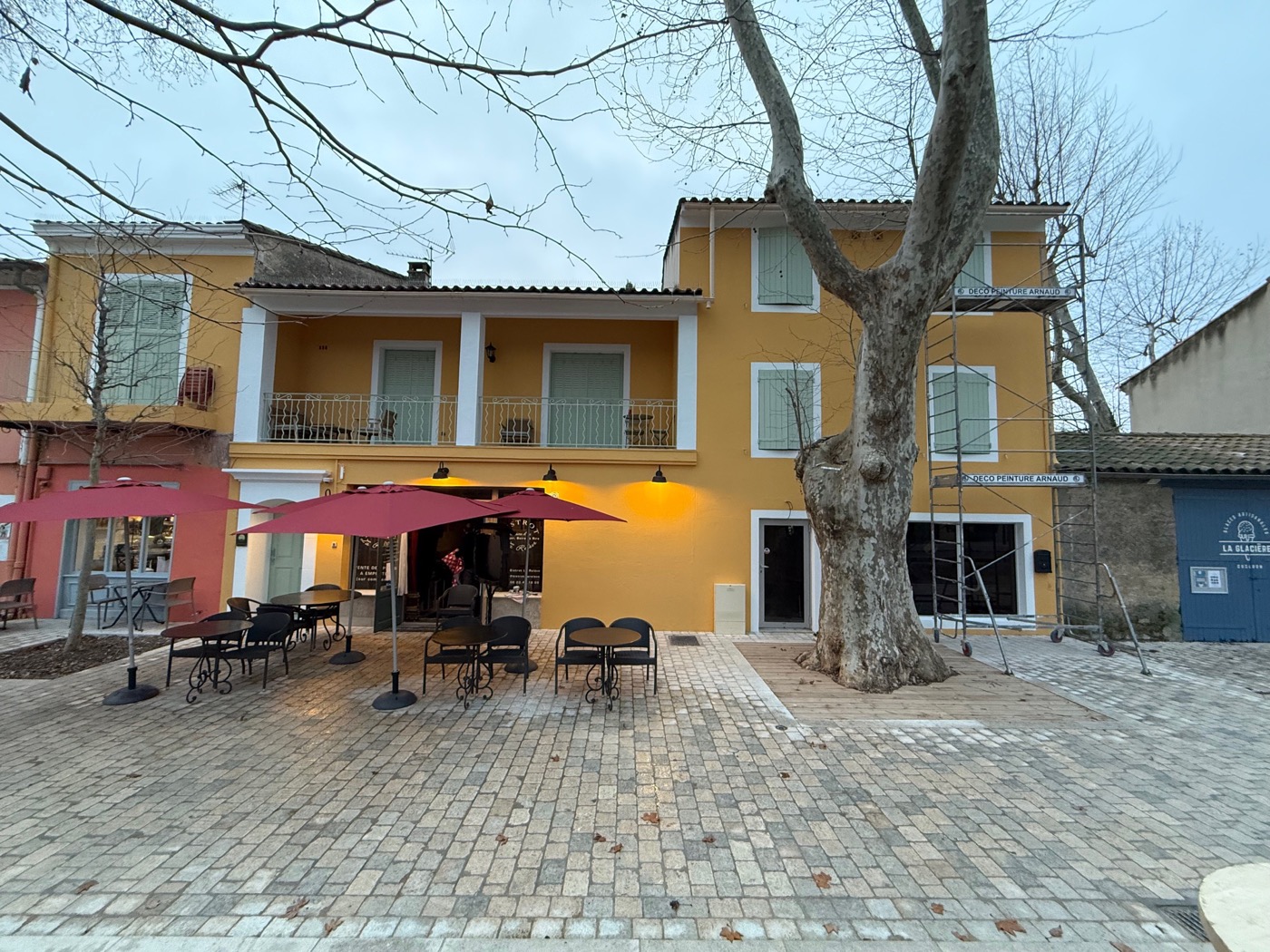 Façade de bistrot de village peinte en jaune ocre, volets vert d'eau et terrasse